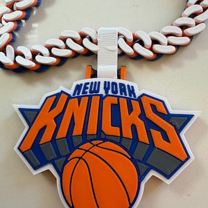 May include: A chunky chain necklace featuring a large pendant with the New York Knicks logo. The chain consists of interlocking white, orange, and blue plastic links. The pendant displays the text New York Knicks in orange and blue over a basketball.