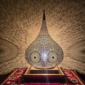 May include: A teardrop-shaped metal table lamp with an intricate, laser-cut filigree pattern that casts detailed, radiating shadows. The lamp features a pointed top, a wide rounded body, and a square base, emitting a warm, bright light.