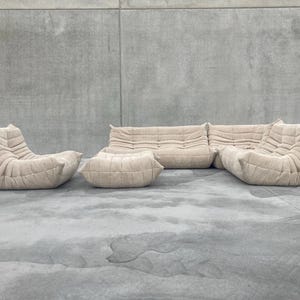 Pode incluir: Conjunto de sof&aacute; modular bege com um design acolchoado texturizado. O conjunto inclui um sof&aacute; de dois lugares, duas poltronas e um pufe. Os m&oacute;veis est&atilde;o dispostos sobre uma superf&iacute;cie de concreto cinza, contra uma parede cinza.
