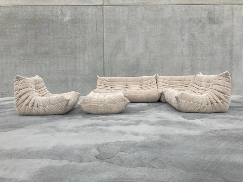 Pode incluir: Conjunto de sof&aacute; modular bege com um design acolchoado texturizado. O conjunto inclui um sof&aacute; de dois lugares, duas poltronas e um pufe. Os m&oacute;veis est&atilde;o dispostos sobre uma superf&iacute;cie de concreto cinza, contra uma parede cinza.