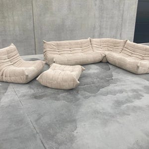 Pode incluir: Um conjunto de sof&aacute;s modulares bege, incluindo um sof&aacute; de canto, uma cadeira individual e um pufe. Os m&oacute;veis t&ecirc;m uma apar&ecirc;ncia macia e texturizada com pregas horizontais. As pe&ccedil;as est&atilde;o dispostas em um piso de concreto.