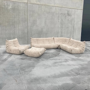 Pode incluir: Um conjunto de sof&aacute;s modular bege, incluindo um sof&aacute; de tr&ecirc;s lugares, duas poltronas e um pufe. Os m&oacute;veis t&ecirc;m um design texturizado e acolchoado. O conjunto est&aacute; colocado sobre uma superf&iacute;cie de concreto, com uma parede cinza e uma porta de garagem cinza escuro ao fundo.