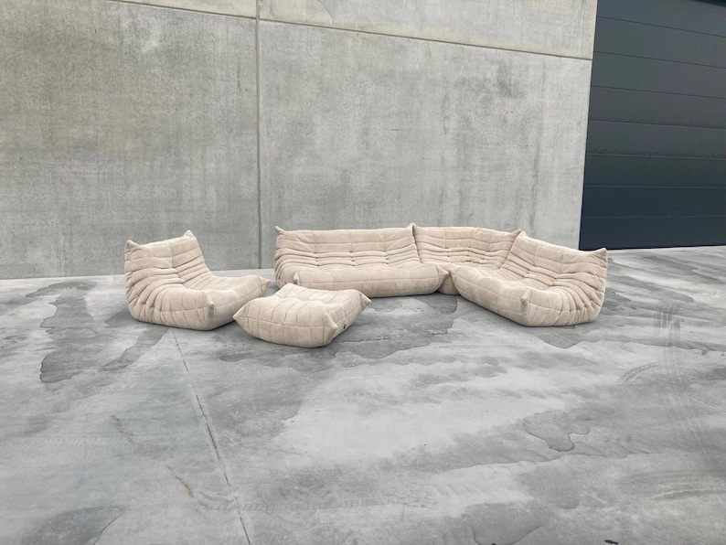 Pode incluir: Um conjunto de sof&aacute;s modular bege, incluindo um sof&aacute; de tr&ecirc;s lugares, duas poltronas e um pufe. Os m&oacute;veis t&ecirc;m um design texturizado e acolchoado. O conjunto est&aacute; colocado sobre uma superf&iacute;cie de concreto, com uma parede cinza e uma porta de garagem cinza escuro ao fundo.
