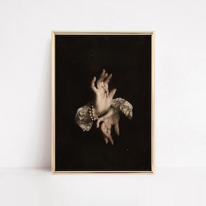 Puede incluir: Una impresión de arte mural enmarcada que presenta una ilustración en tono sepia de dos manos sobre un fondo negro sólido. Las manos están adornadas con una delicada pulsera de perlas y un pequeño colgante de cruz, enmarcadas por puños de encaje.