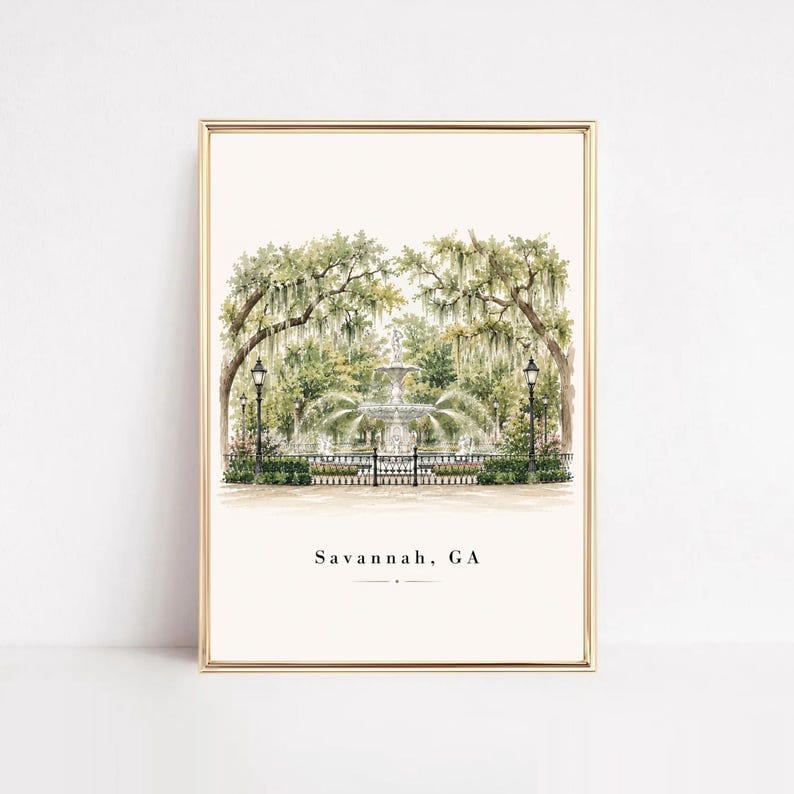 May include: A vertical art print in a gold frame featuring a watercolor illustration of a fountain in a park with large trees draped in Spanish moss. The text Savannah, GA is printed in black at the bottom center of the white background.