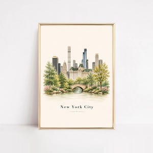 May include: A vertical art print in a gold frame featuring a watercolor illustration of the New York City skyline, a stone bridge over water, and lush green trees. The text New York City is printed in black at the bottom of the cream-colored paper.