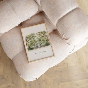 May include: A rectangular framed print featuring a watercolor illustration of a park fountain surrounded by trees with hanging moss. The text Savannah, GA is printed in black at the bottom center of the off-white paper within the light wood frame.