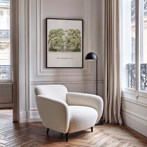 May include: A cream-colored, textured boucl&eacute; armchair with rounded arms and dark wooden legs, positioned near a black floor lamp and a framed wall print featuring a green tree-lined park scene with the text Savannah, GA printed below.