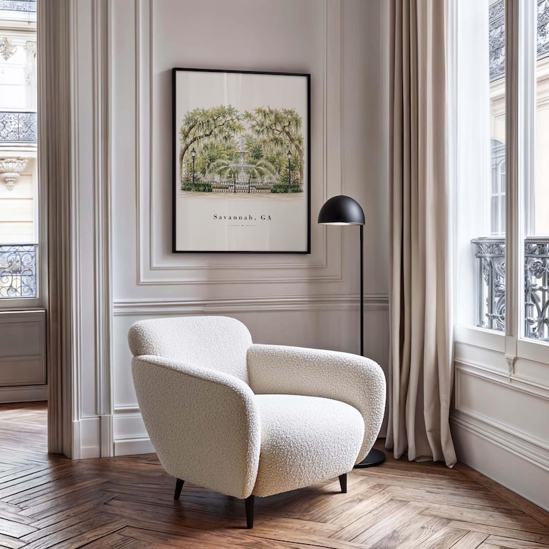 May include: A cream-colored, textured boucl&eacute; armchair with rounded arms and dark wooden legs, positioned near a black floor lamp and a framed wall print featuring a green tree-lined park scene with the text Savannah, GA printed below.