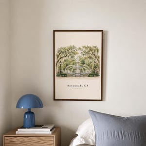 May include: A framed wall art print featuring a watercolor illustration of a fountain in a park with Spanish moss-covered trees and black lampposts. The text Savannah, GA is printed in black at the bottom center of the cream-colored background.