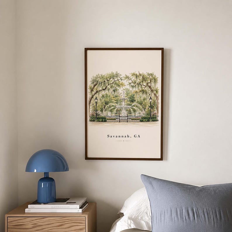 May include: A framed wall art print featuring a watercolor illustration of a fountain in a park with Spanish moss-covered trees and black lampposts. The text Savannah, GA is printed in black at the bottom center of the cream-colored background.