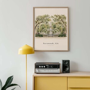 May include: A framed wall art print featuring a watercolor illustration of a fountain in a park with Spanish moss-draped trees. The text Savannah, GA is printed in black at the bottom center of the cream-colored background of the artwork.