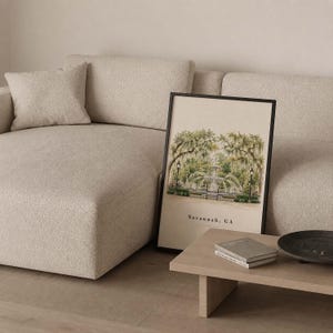 May include: A framed vertical art print featuring a watercolor illustration of a fountain surrounded by oak trees with Spanish moss. The print includes the text Savannah, GA at the bottom, displayed in a black frame against a light beige wall.