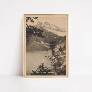 以下が含まれることがあります： 薄いゴールドのフレームに入ったセピア調のヴィンテージ写真プリント。山岳風景、湖、岩の多い海岸線、常緑樹が描かれています。画像の下部には「101. LAKE AGNES」という白い文字が印刷されており、美しい景色を捉えています。