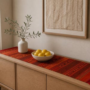 May include: A wooden sideboard with a red and orange woven table runner. A white ceramic vase holds green branches. A white bowl contains several yellow lemons. A framed piece of art hangs on the wall.