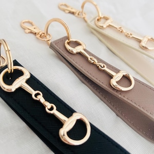 May include: Close-up of three keychains with gold-colored horse bit charms. The keychains have straps in black, taupe, and cream colors. Each keychain has a gold-colored clasp and ring.