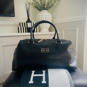 May include: Black leather duffel bag with gold-tone hardware and handles, placed on a black and white patterned blanket. The scene includes a decorative vase with greenery. The bag has a gold emblem.