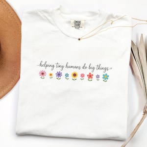 Peut inclure: Un t-shirt blanc à manches courtes avec le texte noir "helping tiny humans do big things" imprimé sur la poitrine. Sous le texte se trouve une rangée de neuf petites fleurs illustrées colorées dans des tons rose, jaune, violet, bleu et orange.