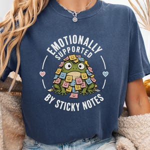 May include: A navy blue crew neck t-shirt featuring a graphic of a green frog covered in colourful sticky notes with smiley faces. The text reads "EMOTIONALLY SUPPORTED BY STICKY NOTES" in white capital letters, flanked by two small hearts.