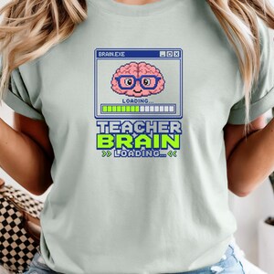 May include: A light sage green t-shirt featuring a pixel art graphic of a pink brain wearing blue glasses inside a computer window labelled BRAIN.EXE. Below the window, the text reads TEACHER BRAIN LOADING with a green progress bar.