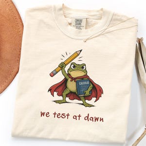 May include: A cream-coloured t-shirt featuring a graphic of a green frog wearing a red cape, holding a yellow pencil and a blue book labelled Calculus. Below the frog, the text reads we test at dawn. The shirt has a Comfort Colors brand neck label.