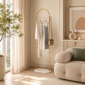 Puede incluir: Un perchero de metal dorado con una parte superior arqueada y una base ovalada blanca. El perchero cuenta con una barra horizontal para colgar ropa, que sostiene una prenda blanca y una azul claro, con un pequeño bolso de color canela a un lado.