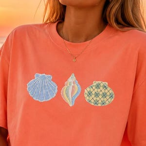 Peut inclure: Un t-shirt à manches courtes de couleur corail avec une impression graphique de trois coquillages sur la poitrine. Le coquillage de gauche a un motif vichy bleu, celui du centre est aux tons pastel et celui de droite a un motif écossais vert et jaune.