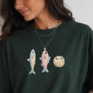 Puede incluir: Una camiseta de manga corta en color verde bosque oscuro con un estampado gráfico de tres peces pequeños en tonos azul claro, rosa y amarillo. La prenda tiene cuello redondo y un corte holgado, combinada con pantalones cortos deportivos negros.