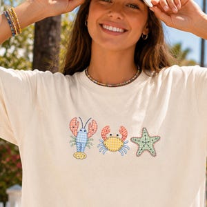 Könnte beinhalten: Ein cremefarbenes Kurzarm-T-Shirt mit einer zentralen Stickerei, die einen blauen Hummer, eine gelbe Krabbe und einen grünen Seestern zeigt. Das T-Shirt wird mit einer beigen Baseballkappe, weißen Shorts, einer Perlenkette und Perlenarmbändern kombiniert.
