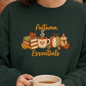 Könnte beinhalten: Ein dunkelwaldgrünes Sweatshirt mit Rundhalsausschnitt und einem Grafikdruck, der einen Bücherstapel, eine Kaffeetasse mit Herzmotiv, einen Kürbis und eine Kerze zeigt, sowie dem Schriftzug "Autumn Essentials" in goldgelber Schreibschrift.