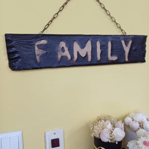 May include: A wooden sign with the word "FAMILY" carved into it. The sign is painted dark blue and hangs from a metal chain. The letters are a lighter wood color, creating a contrast.