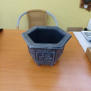 Può includere: Un vaso esagonale grigio scuro con una finitura usurata. L'esterno presenta motivi decorativi in rilievo e il carattere cinese per la buona fortuna, 福, centrato sul pannello frontale. Il vaso ha un bordo largo e piccoli piedini.