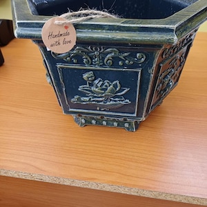 Può includere: Un vaso esagonale verde scuro con una finitura anticata e motivi floreali in rilievo su ogni lato. Un'etichetta rotonda in carta marrone è fissata con dello spago e riporta il testo "Handmade with love" e una piccola icona a cuore rosso.