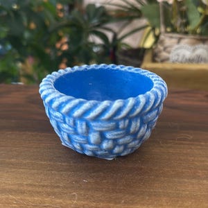 Op de afbeelding: Een kleine, ronde, blauwe kom met een getextureerde buitenkant die het uiterlijk van een gevlochten mand nabootst. De rand heeft een opvallend touwachtig patroon en de blauwe kleur is geaccentueerd met een lichtere, verweerde witte afwerking.