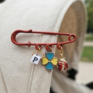 Può includere: Una spilla da balia rossa con tre ciondoli pendenti: una perlina cubica bianca con la lettera P nera, un ciondolo a forma di quadrifoglio con smalto giallo e blu e una piccola coccinella rossa e nera con dettagli in cristallo trasparente.