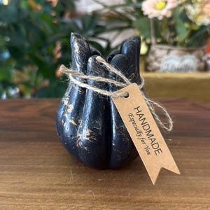 May include: A black, bulbous, multi-lobed decorative object with gold-colored veining. It is wrapped in natural twine with a light brown, rectangular paper tag attached that reads: HANDMADE Especially for You in black, sans-serif lettering.