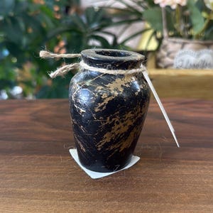 May include: A small, rounded vase with a black base color and distressed gold-toned paint accents. The neck of the vase is wrapped with a thin, natural-colored jute twine, and a small white paper tag is attached to the twine on the right side.