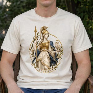 May include: A cream-colored short-sleeve t-shirt featuring a graphic print of the Greek goddess Athena. She wears a golden helmet and breastplate, holds a spear, and carries an owl. The design is framed by olive branches and the text ATHENA.
