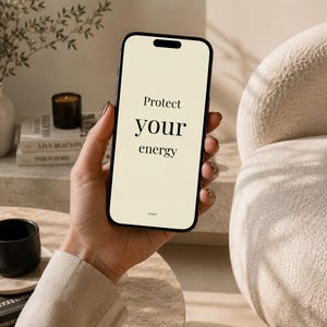 Könnte beinhalten: Ein schwarzes Smartphone zeigt den Text "Protect your energy" auf einem cremefarbenen Bildschirm. Das Telefon wird in einer Hand gehalten. Eine kleine schwarze Tasse und eine Kerze sind im Hintergrund zu sehen.