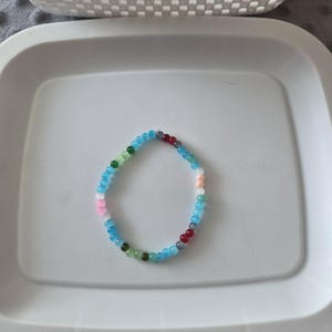 May include: A circular bracelet composed of small, smooth, translucent beads in a variety of colors including light blue, dark blue, green, red, pink, white, and peach. The beads are strung together in a continuous loop, creating a simple design.