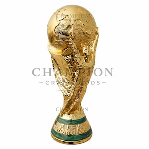 May include: A gold-colored trophy with a globe on top, held by two figures. The base is green with gold stripes and the words "FIFA WORLD CUP". The trophy is a symbol of victory.