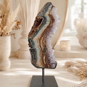 Könnte beinhalten: Eine vertikale Scheibe einer Achat- und Amethyst-Kristallgeode auf einem schwarzen Metallständer. Der Stein zeigt konzentrische Bänder in Braun, Beige, Weiß und Grau mit einem gezackten, kristallinen violetten Amethystrand an einer Seite.