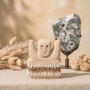 Könnte beinhalten: Eine beigefarbene Steinskulptur einer Figur mit erhobenen Armen, verziert mit einem Band aus weißen Muscheln und Bindfaden. Im Hintergrund befindet sich eine große, gemusterte Geode auf einem Ständer, zusammen mit getrockneten Blumen, die eine natürliche, erdige Ästhetik schaffen.
