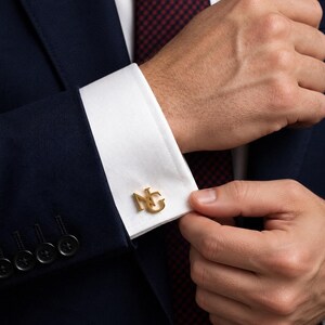 Op de afbeelding: Een paar goudkleurige metalen manchetknopen met de in elkaar grijpende letters N en G. De manchetknopen zijn bevestigd aan de witte manchet van een overhemd met lange mouwen, gedragen onder een donkerblauw colbert met vier knopen op de mouw.