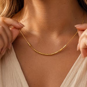 以下が含まれることがあります： ゴールドトーンのネックレスで、繊細なチェーンの中央に小さな円筒形と球形のビーズが直線状に並んでおり、鎖骨に沿って配置された控えめで質感のあるデザインが特徴です。