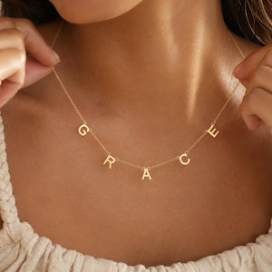 Pode incluir: Um colar de tom dourado com uma corrente delicada e cinco letras maiúsculas individuais suspensas ao longo da corrente, soletrando o nome GRACE numa fonte sem serifa limpa. As letras estão espaçadas uniformemente na clavícula.