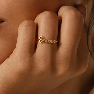 May include: A gold-toned ring featuring the name Emma in a cursive script font. The ring is worn on a finger, showcasing the polished metal finish and the delicate, flowing lettering of the name design across the top of the band.