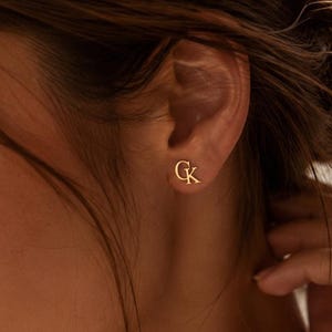 Puede incluir: Un pequeño pendiente de botón dorado con las letras G y K unidas. El pendiente tiene un acabado metálico pulido y se lleva en el lóbulo de la oreja. El diseño consiste en una letra G mayúscula situada junto a una letra K mayúscula.