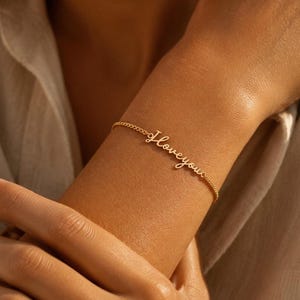 Könnte beinhalten: Ein goldfarbenes Kettenarmband mit einem zarten Metallanhänger in Schreibschrift, der die Aufschrift I love you trägt. Das Armband besteht aus einer feinen Gliederkette, die das Handgelenk umschließt, wobei der kursive Schriftzug mittig integriert ist.