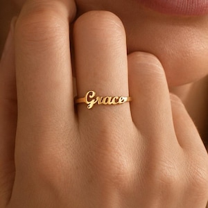 Puede incluir: Un anillo de tono dorado que presenta el nombre Grace en una fuente de escritura cursiva. El anillo consiste en una banda de metal delgada y pulida que se conecta a las letras, las cuales se sitúan horizontalmente sobre el dedo. El metal es liso.